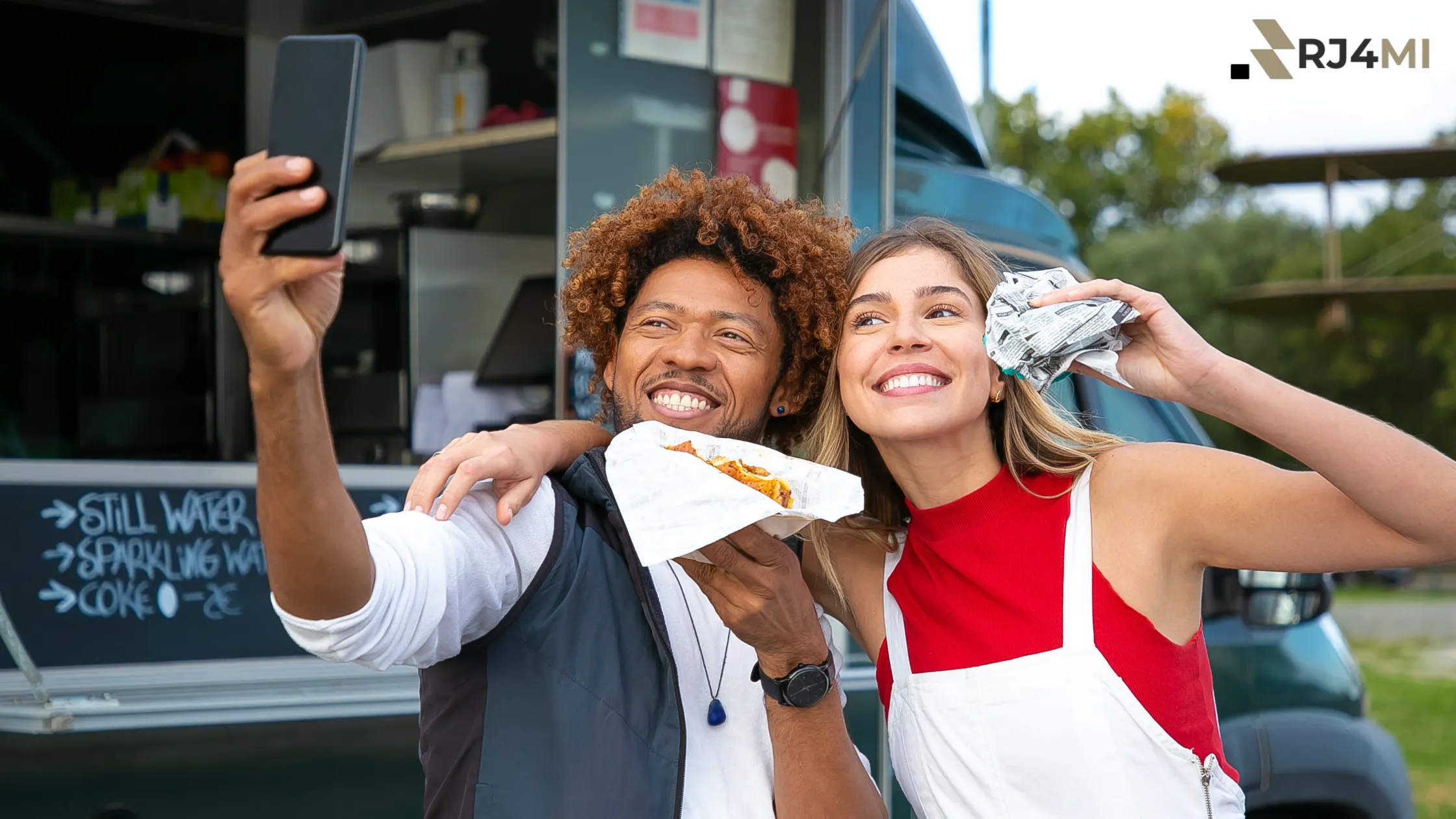 A smiling couple enjoying food from a food truck while taking a selfie at an event.